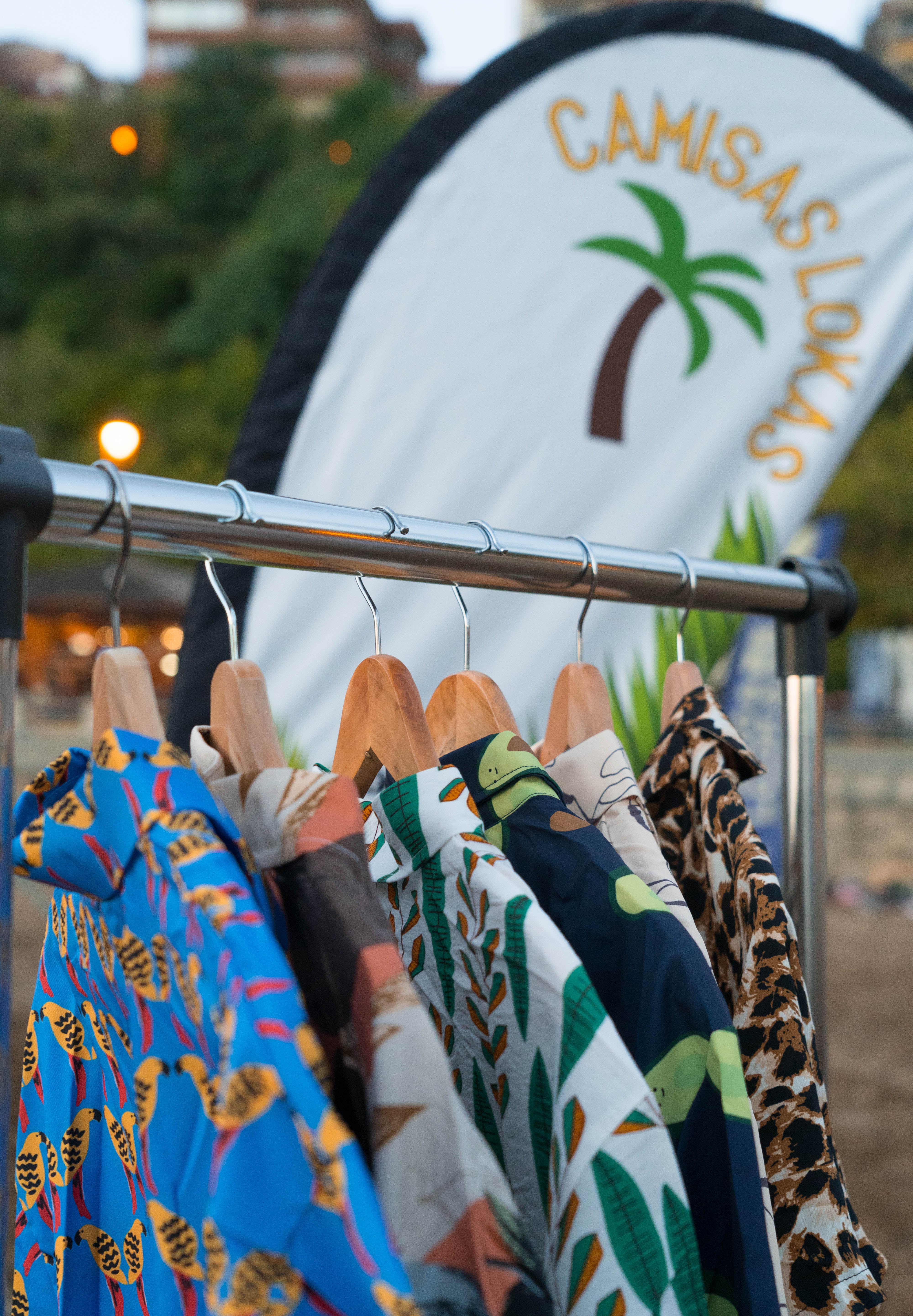 A clothing rack displaying a variety of colorful patterned shirts, including a blue butterfly print, a green palm tree design, and a leopard print shirt, with a white curved banner in the background reading 'CAMISAS LOKAS' and featuring a palm tree graphic.