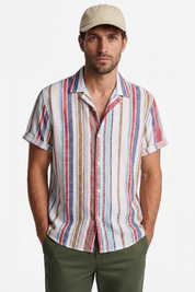 A man wearing a short-sleeve button-up shirt with vertical multicolor stripes in red, blue, beige, and white, paired with a beige baseball cap and olive green pants, against a plain white background.